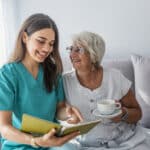 Happy patient is holding caregiver for a hand while spending time together. Elderly woman in nursing home and nurse. Aged elegant woman and tea time at nursing home
